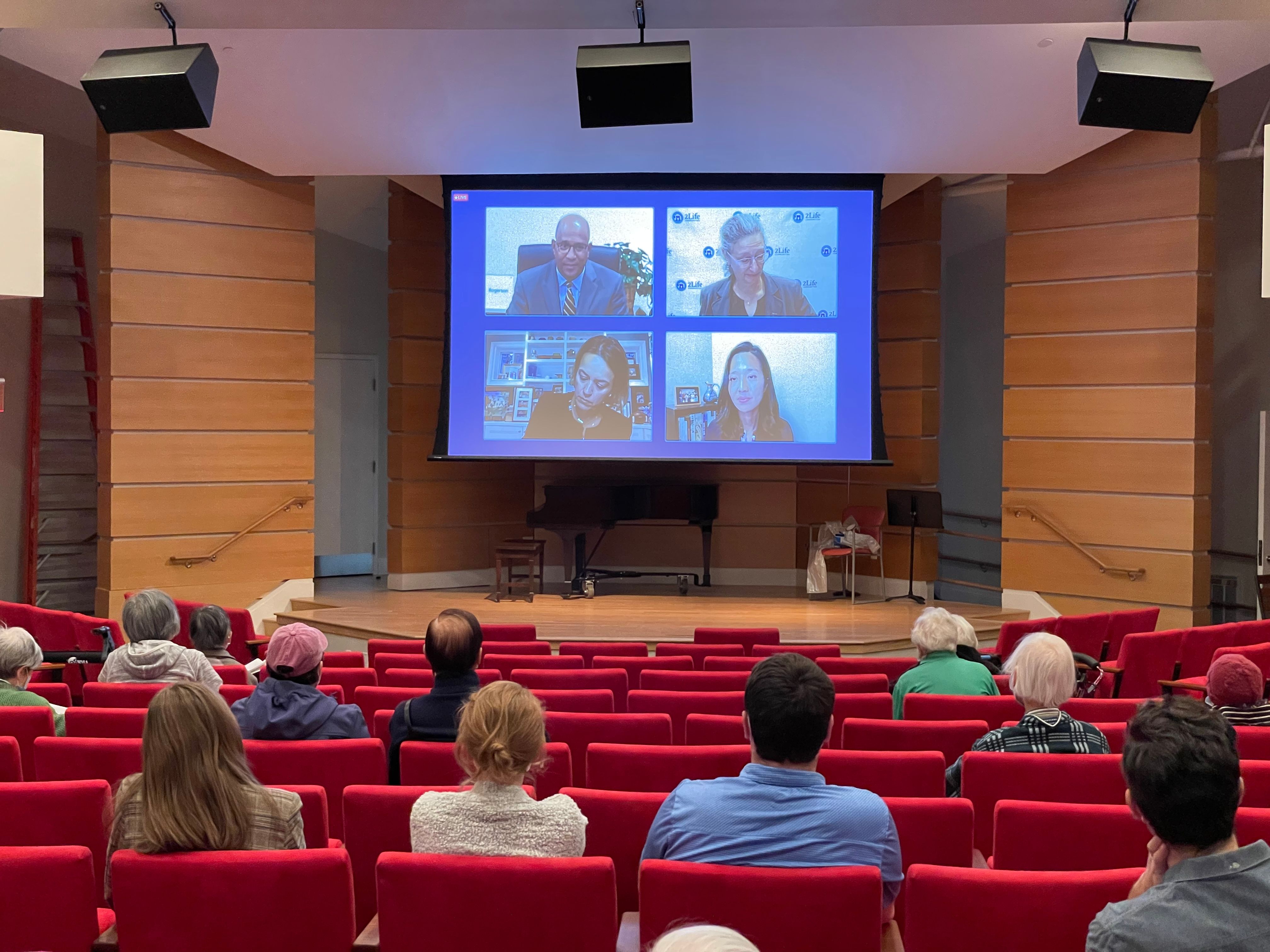 Walter Ramos, Amy Schectman, Annissa Essaibi-George, and Michelle Wu on screen in 2Life's Brighton Campus auditorium during the live broadcast of Candidate Forum: A Vision for Boston Seniors