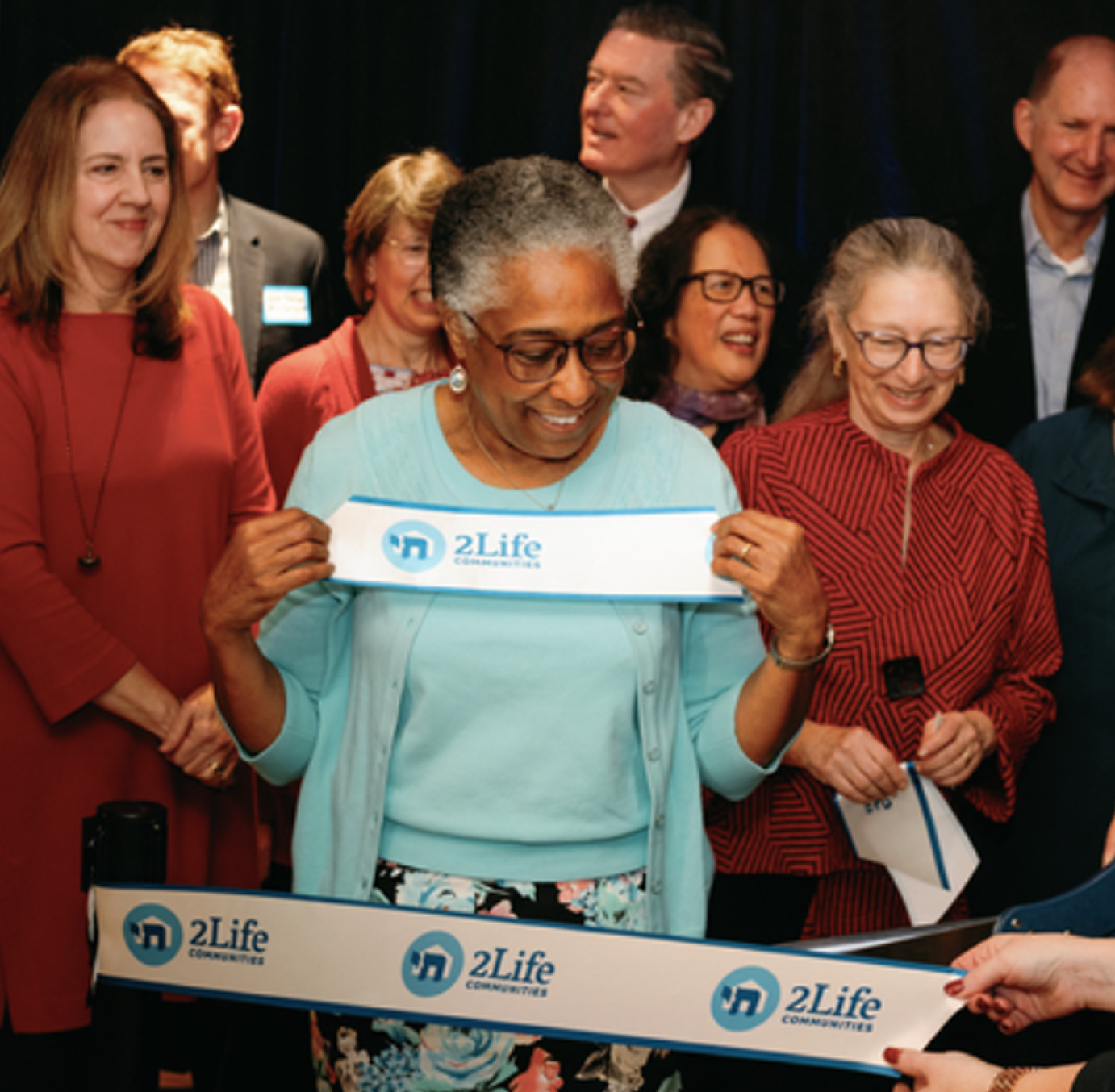 A group of adults at a ribbon-cutting event, with a woman holding the ribbon marked “2Life.”