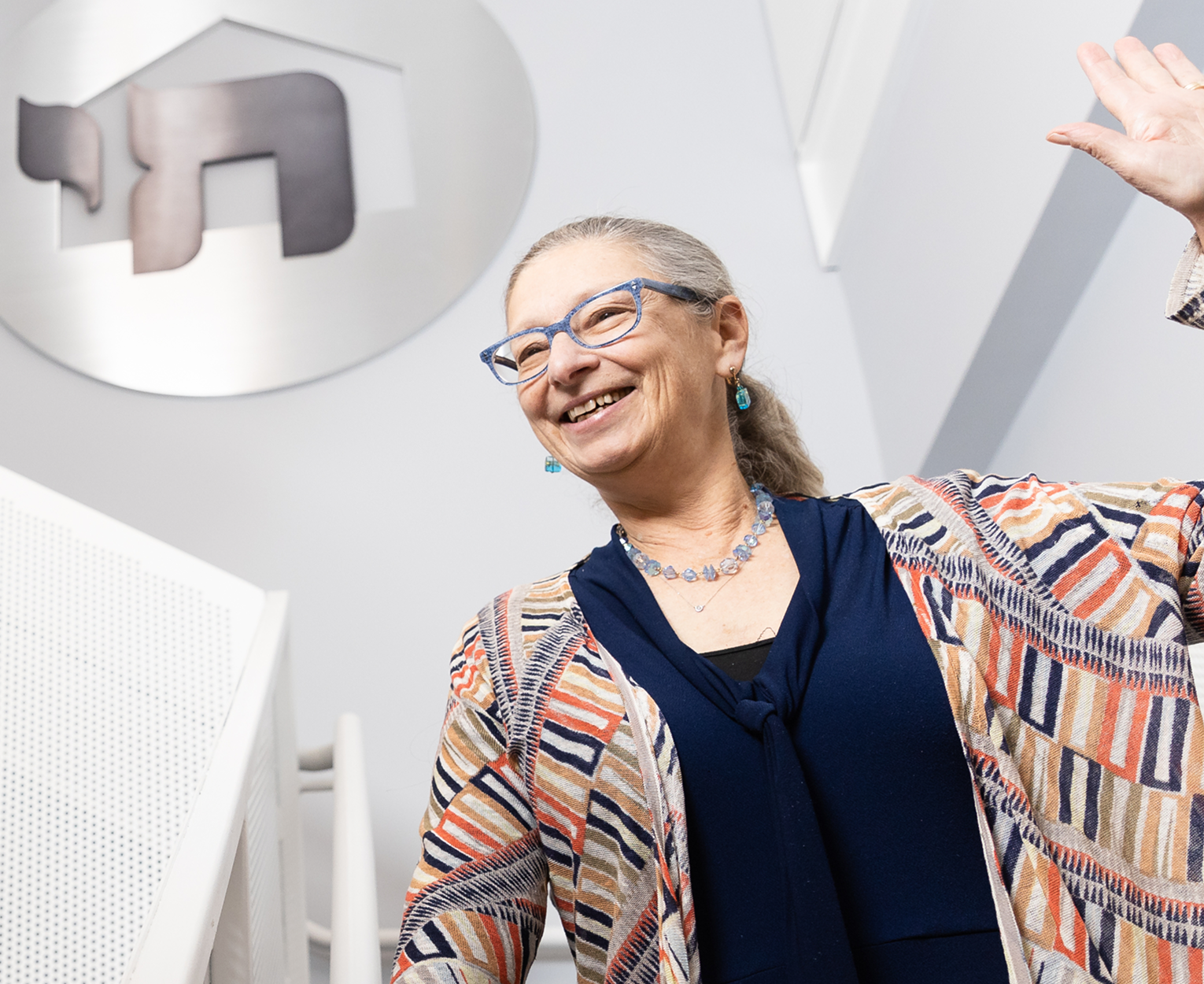 A smiling woman waves while standing indoors near a large decorative sign.