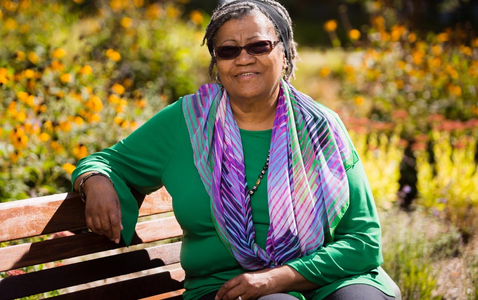 A Black woman sitting on a bench wearing sunglasses, a green shirt and a purple scarf
