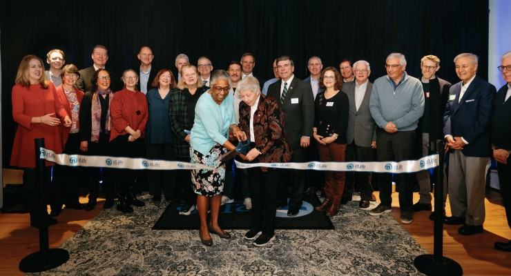 Ribbon-cutting ceremony at a 2Life Communities event, featuring two women cutting the ribbon while surrounded by a diverse group of attendees in formal and business casual attire. The event takes place indoors with a dark curtain backdrop and branded ribbon