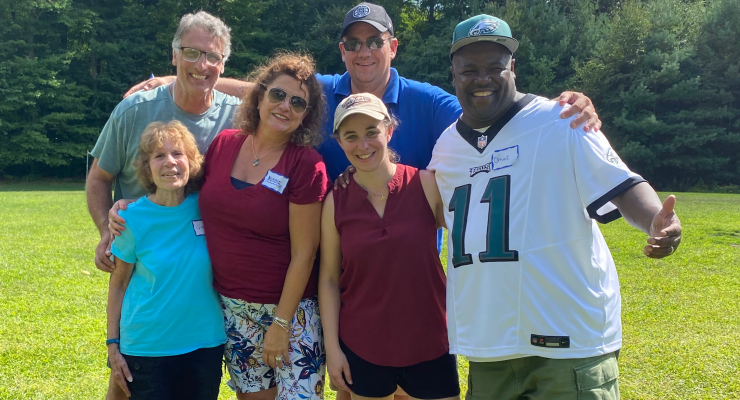 Six smiling people of various ages and backgrounds pose for a group photo on a grassy field with trees in the background.