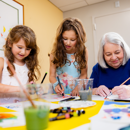 Two young girls and a senior woman painting together at a table.