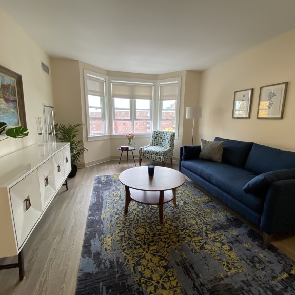 A living room with a couch, dresser, table, rug and window