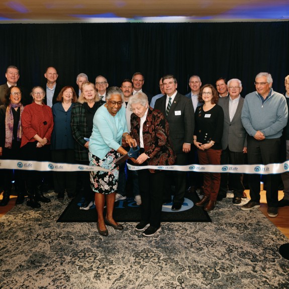 Ribbon-cutting ceremony at a 2Life Communities event, featuring two women cutting the ribbon while surrounded by a diverse group of attendees in formal and business casual attire. The event takes place indoors with a dark curtain backdrop and branded ribbon