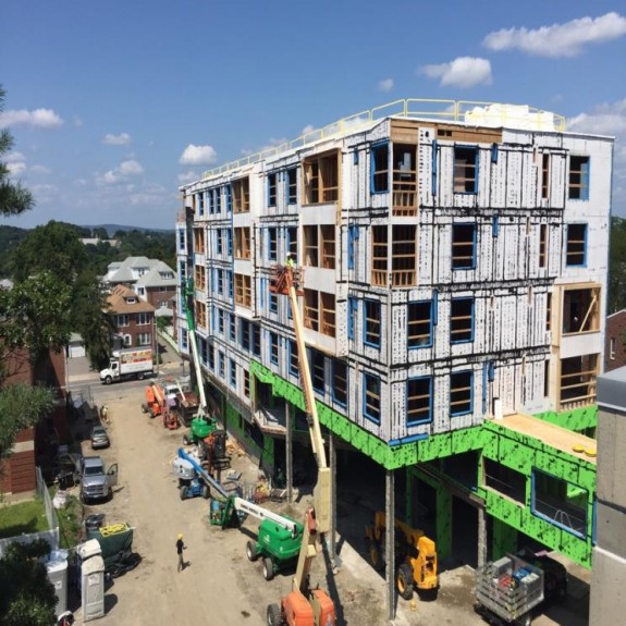 Multi-story building under construction with exposed wooden framing and insulation panels. Construction workers use aerial lifts and heavy machinery to work on the exterior. The site is active with equipment, vehicles, and workers on a dirt road, with a residential neighborhood and rolling hills in the background