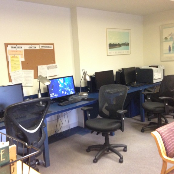 Small computer lab with a row of desktop computers on a blue desk, ergonomic office chairs, and task lamps. A cork bulletin board with posted notices is on the wall, along with framed artwork. A bookshelf with books and a cushioned chair are in the foreground, giving the space a library-like feel.