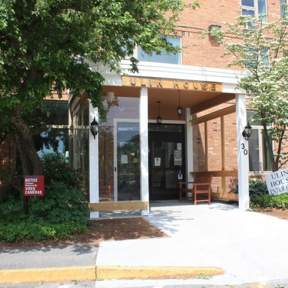 Entrance of Ulin House, a senior living community, featuring a modern glass doorway with a white overhang and signage. The brick building is surrounded by lush greenery and flowering trees. A wooden bench sits near the entrance, and a sign directs visitors to the entryway. The area is well-maintained with accessible pathways.