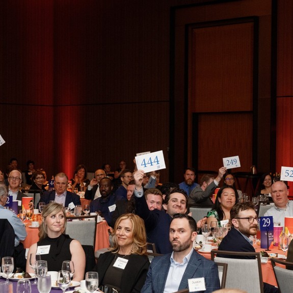 Gala dinner attendees actively participating in an auction, holding up bid cards in a large event space. Fundraising, community event.