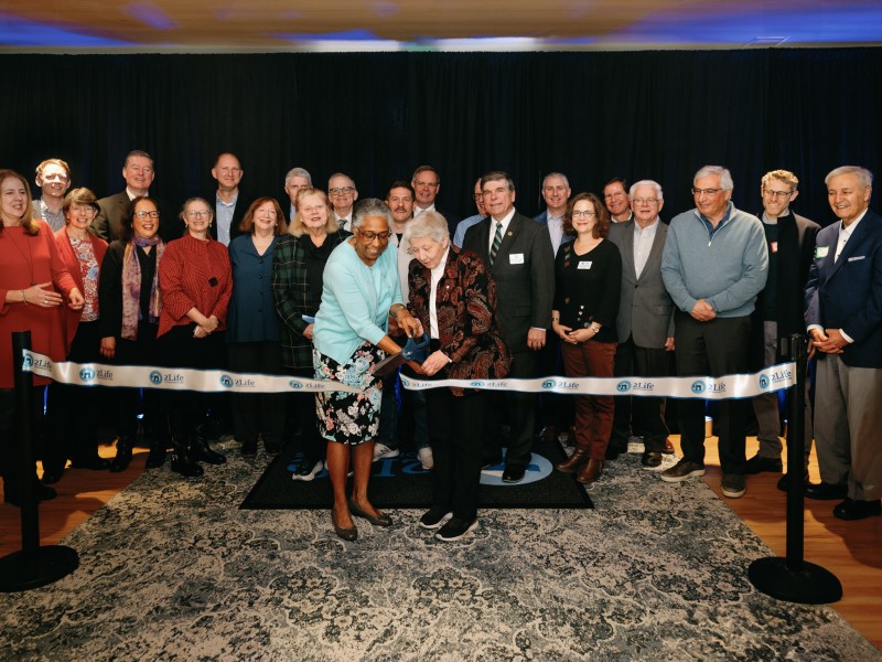 Ribbon-cutting ceremony at a 2Life Communities event, featuring two women cutting the ribbon while surrounded by a diverse group of attendees in formal and business casual attire. The event takes place indoors with a dark curtain backdrop and branded ribbon