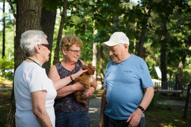 Three people standing in a wooded area; one is holding a small brown dog