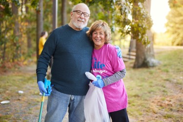 Two people hugging in a park; one holding a grabbing tool, one with a trash bag