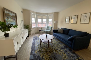 A living room with a couch, dresser, table, rug and window