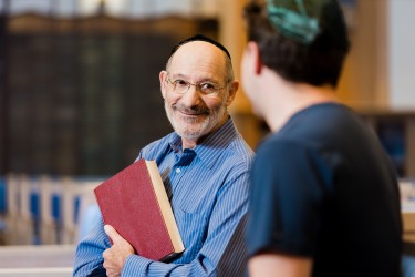 A man holding a book and wearing a yarmulke smiles in conversation with another person in a yarmulke