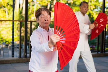 An Asian person wearing a white shirt holds an opened red fan in front of them
