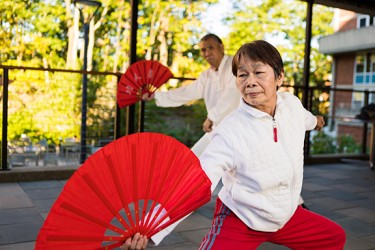 An Asian person wearing a white shirt and red pants crouches while holding out a red fan