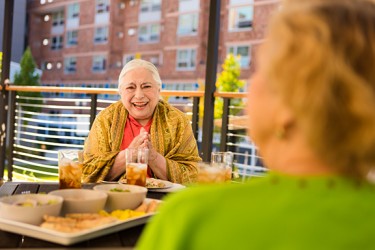 A laughing older person sits at a table during a meal with their hands together
