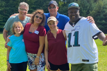 Six smiling people of various ages and backgrounds pose for a group photo on a grassy field with trees in the background.
